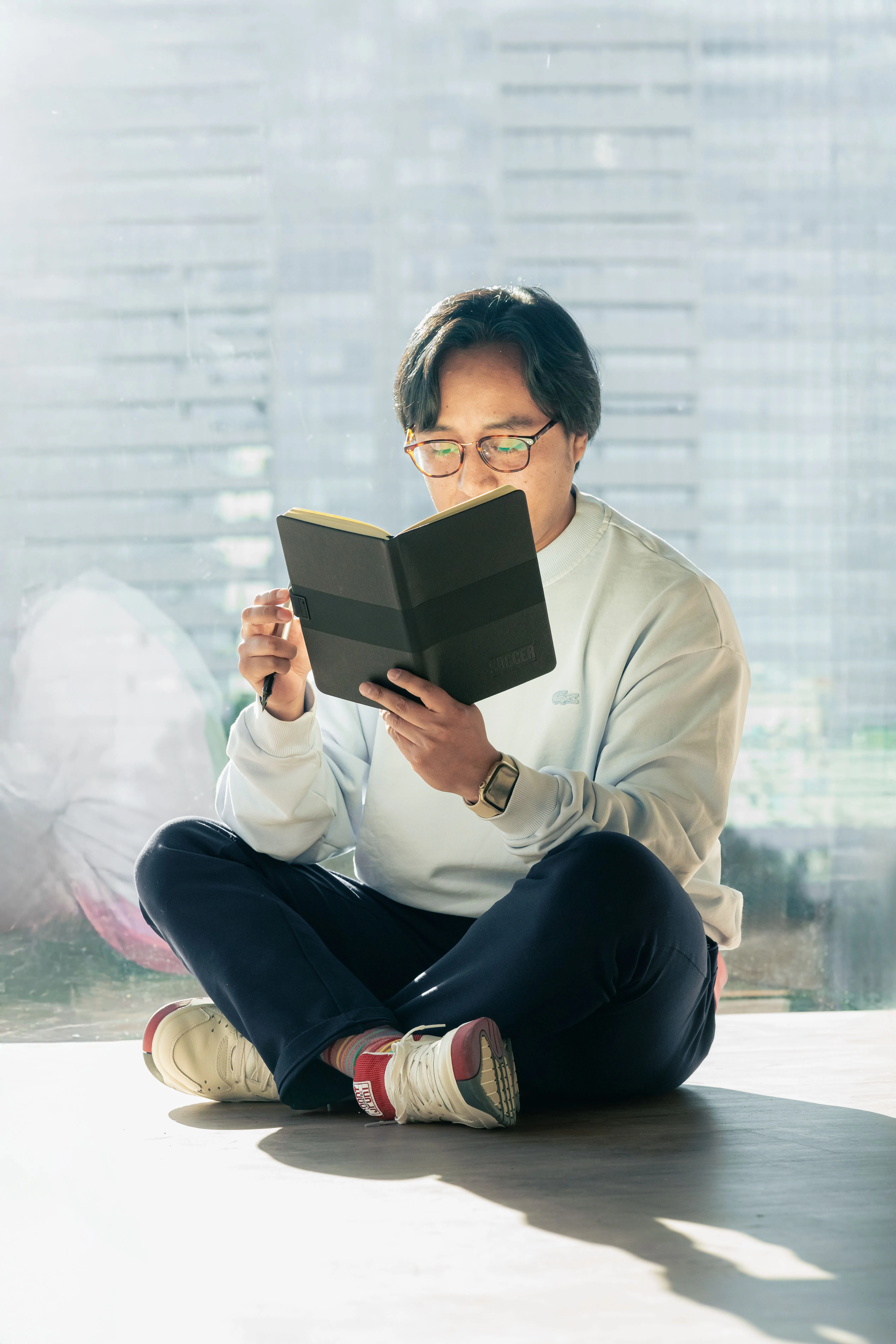 A man in casual attire sits cross-legged, engrossed in a book by the window.
