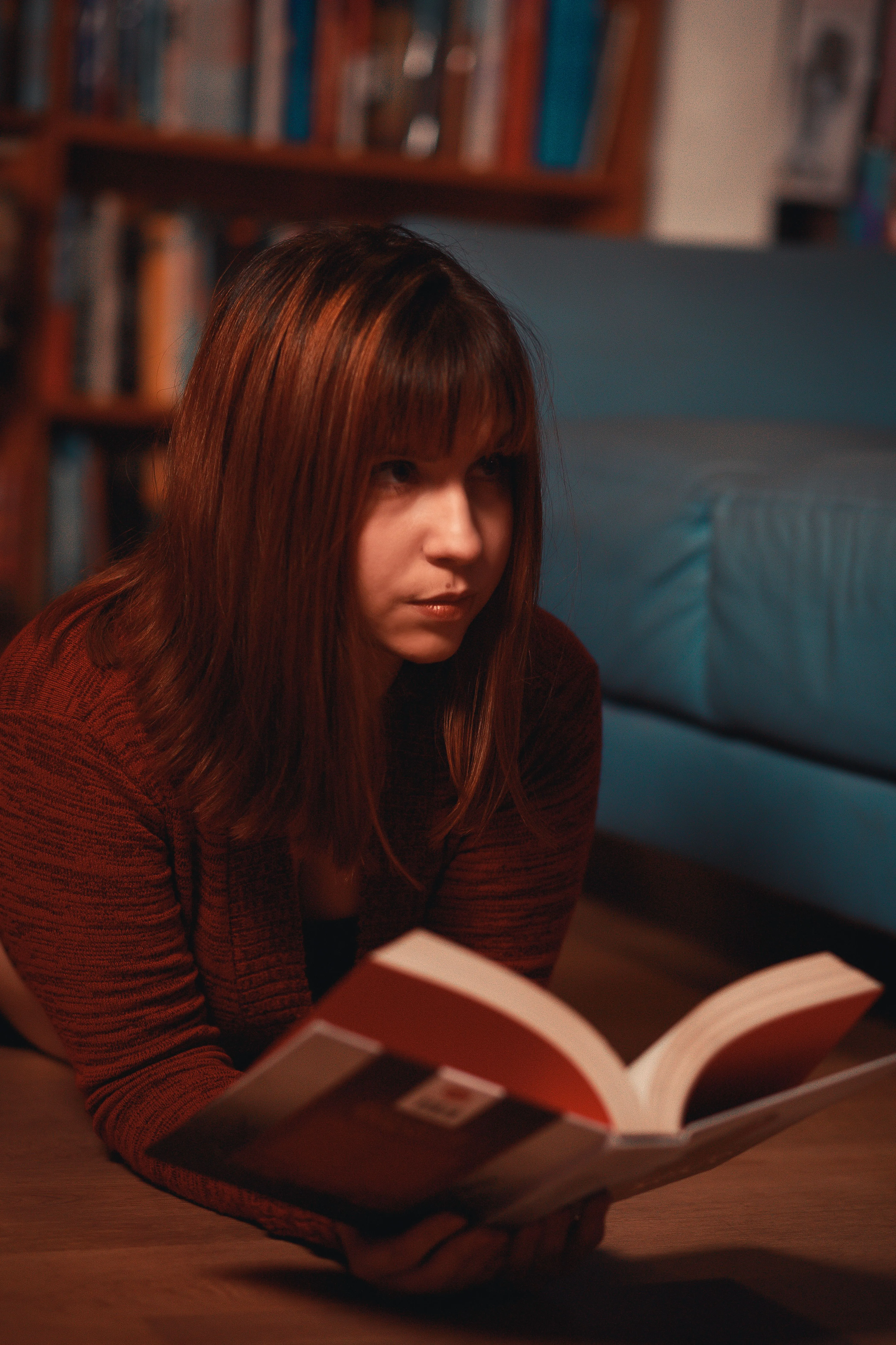 Young woman reading on the floor, surrounded by bookshelves. Comfortable indoor ambiance.