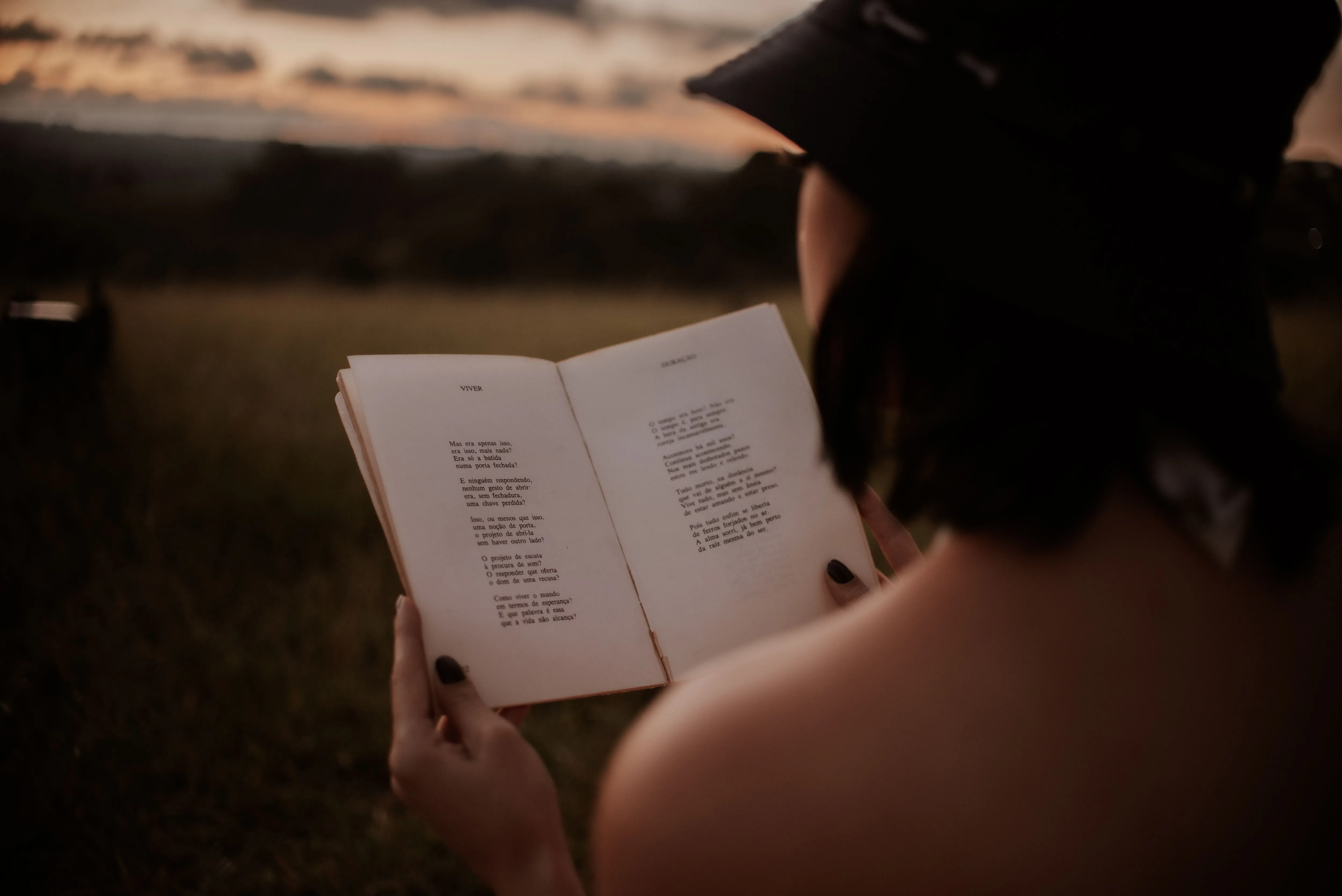 Back view of anonymous female in hat reading book with poetry in grassy field on blurred background on summer evening