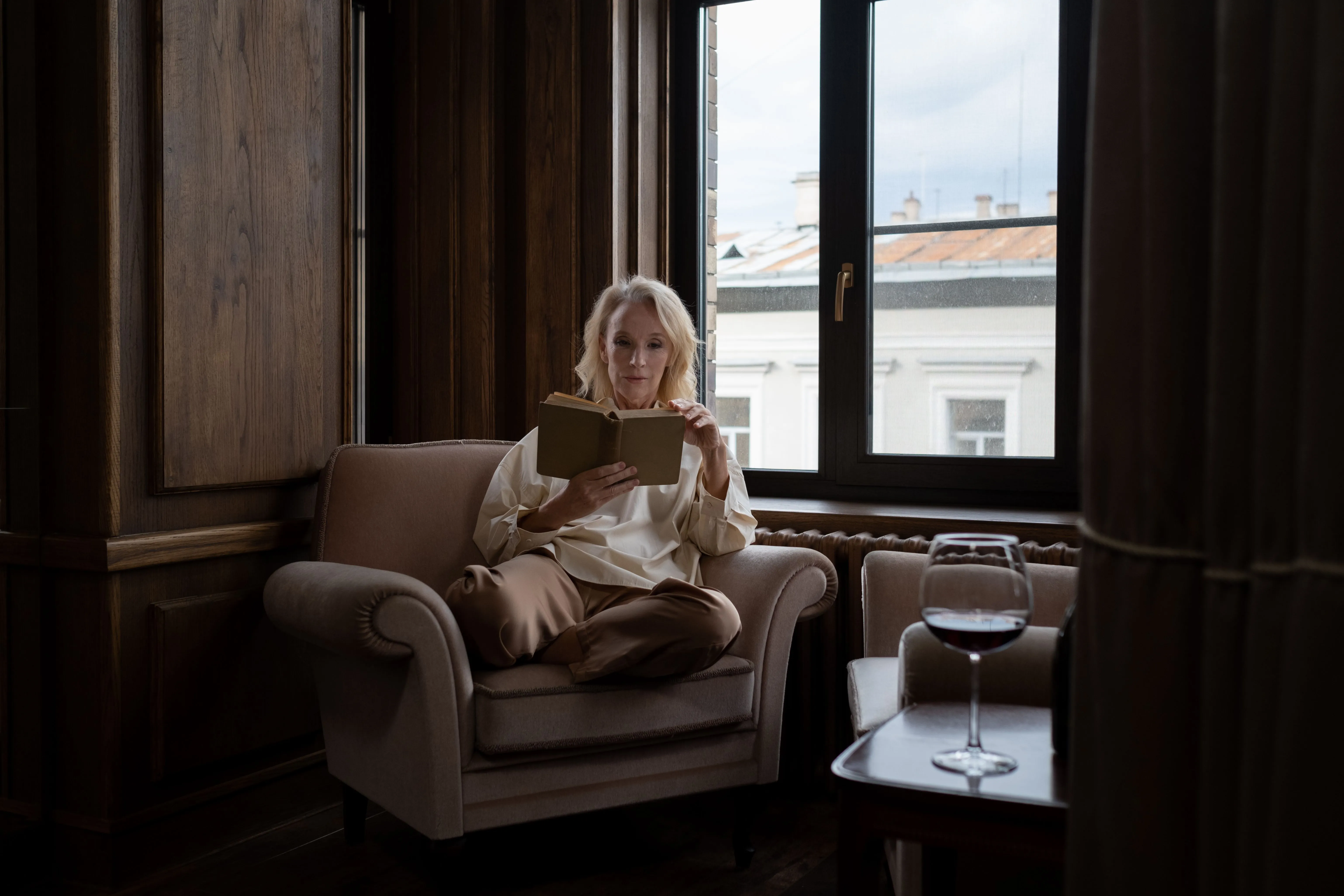 A woman enjoys a book in a cozy living room with a glass of wine by the window.