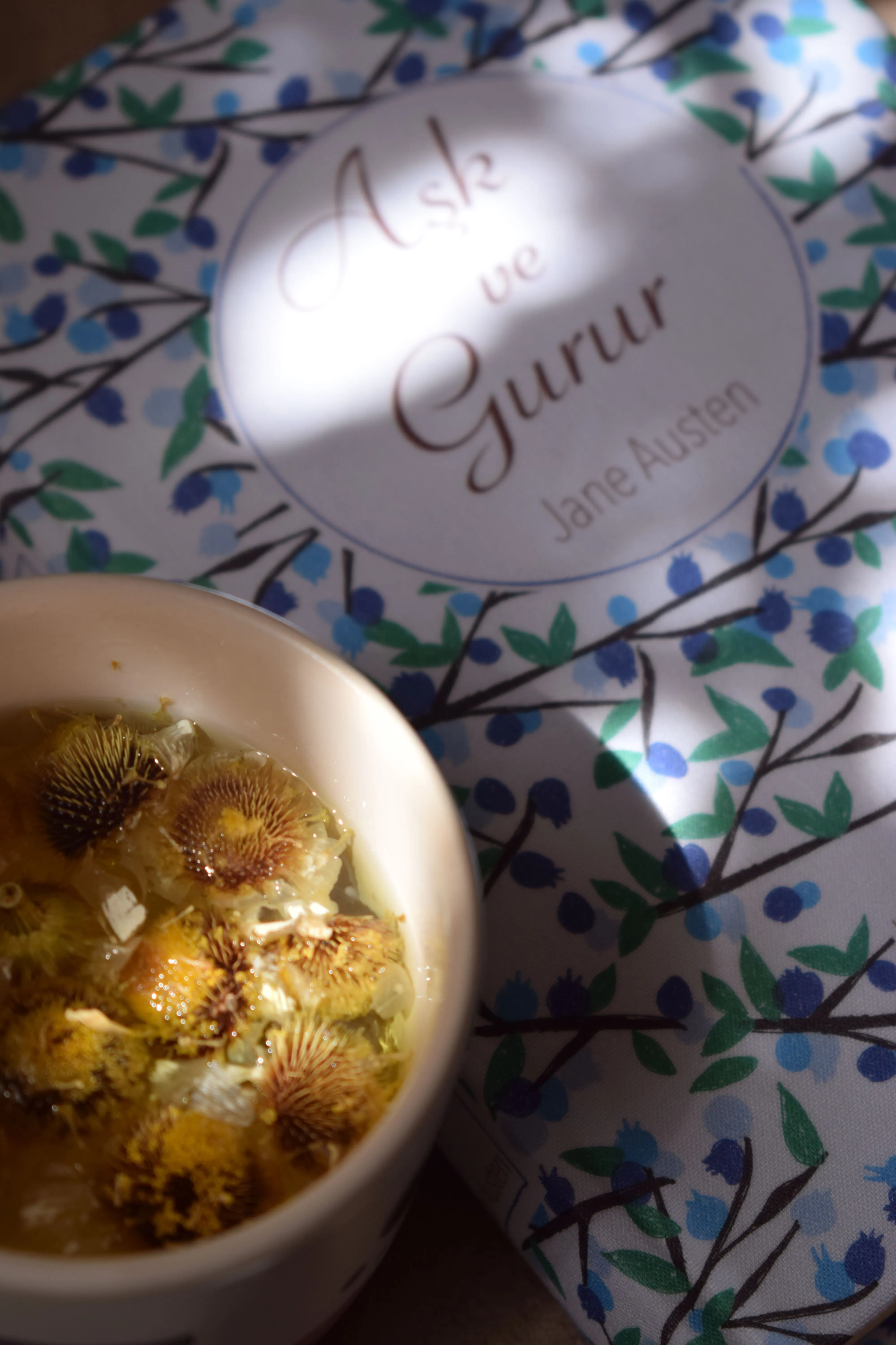 Cozy scene with herbal tea next to a book cover featuring floral patterns in sunlight.