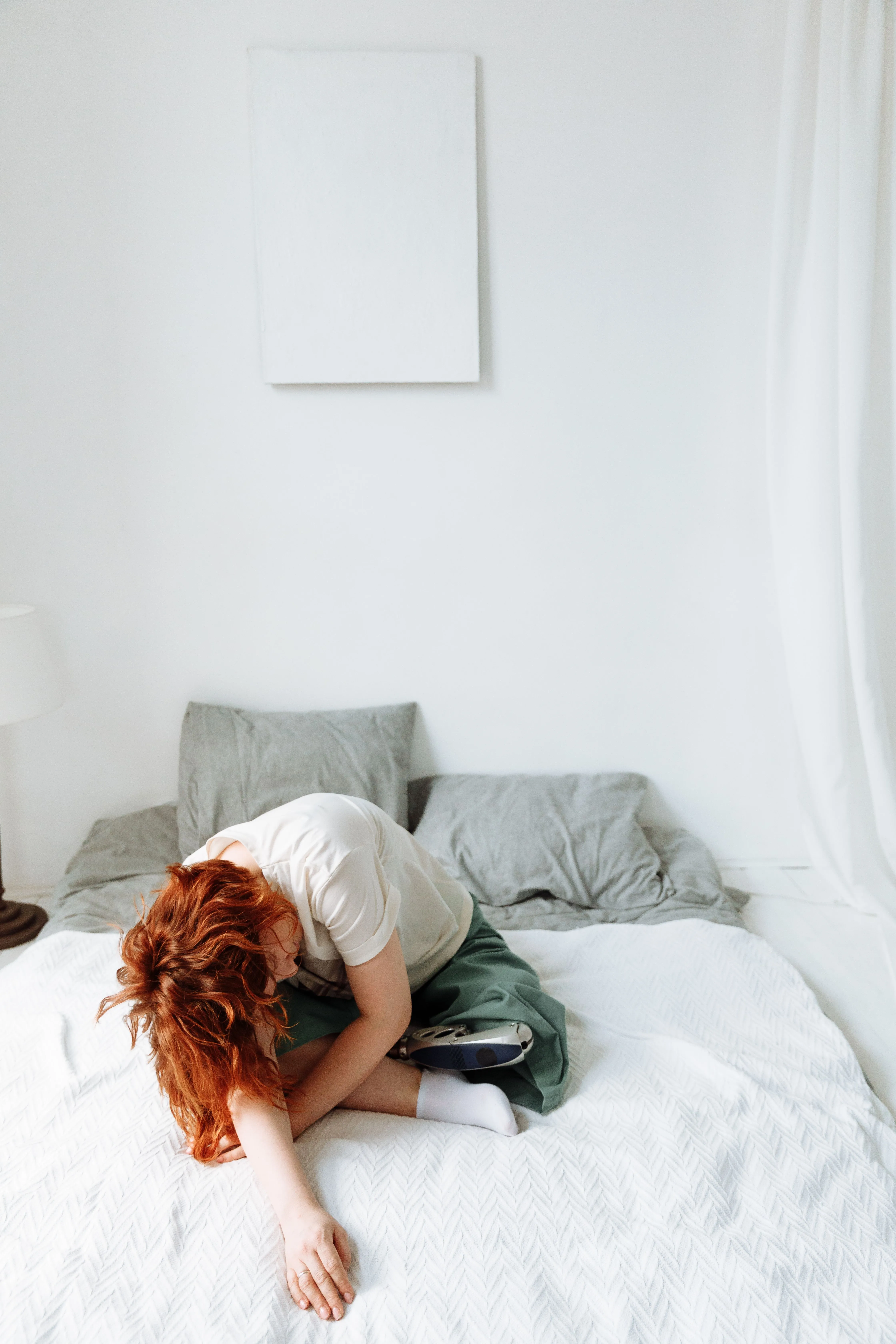 A woman with red hair sitting on a bed in a bright, minimalist bedroom, reflecting solitude and contemplation.