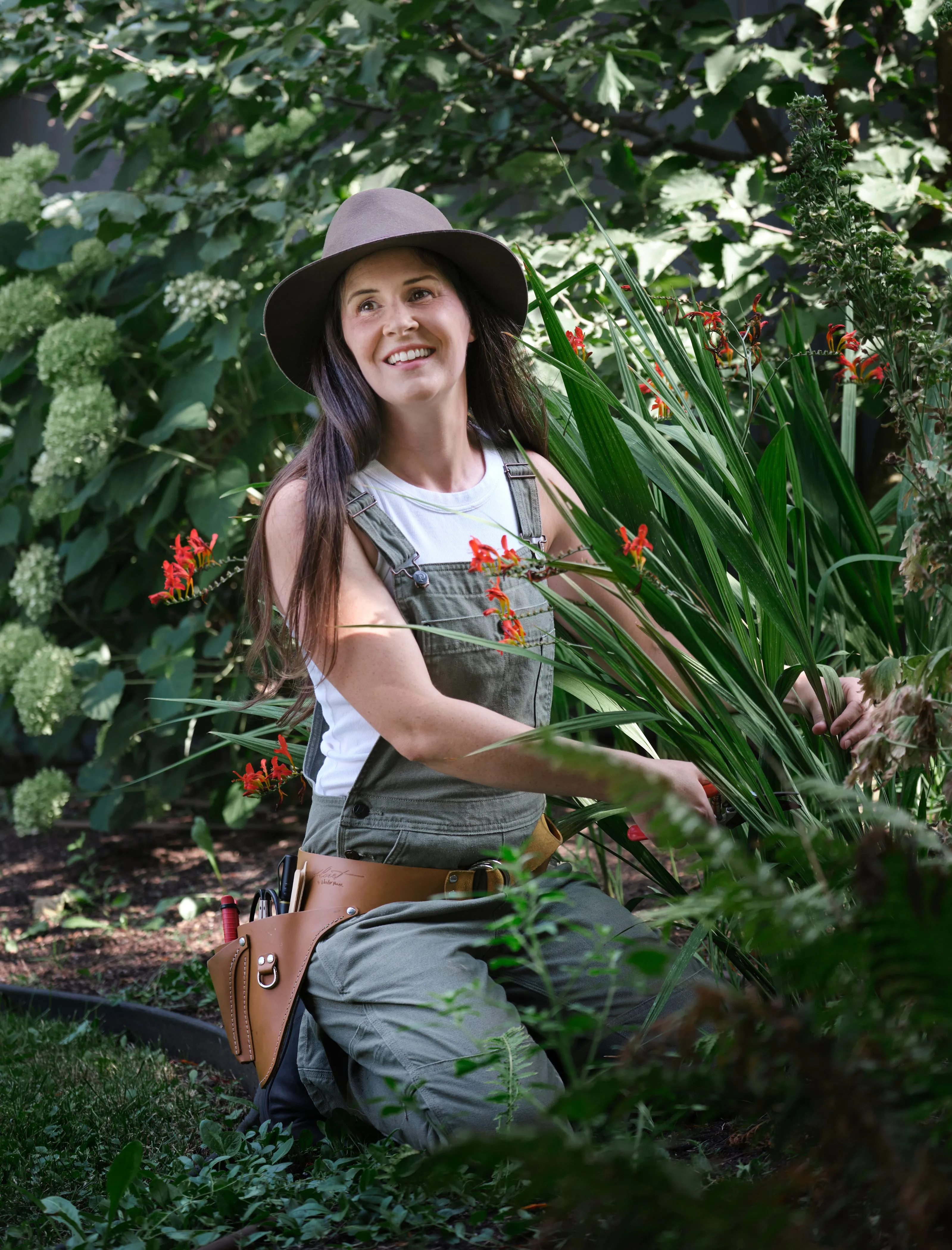 A woman in overalls and a hat tends to vibrant flowers in a lush summer garden.