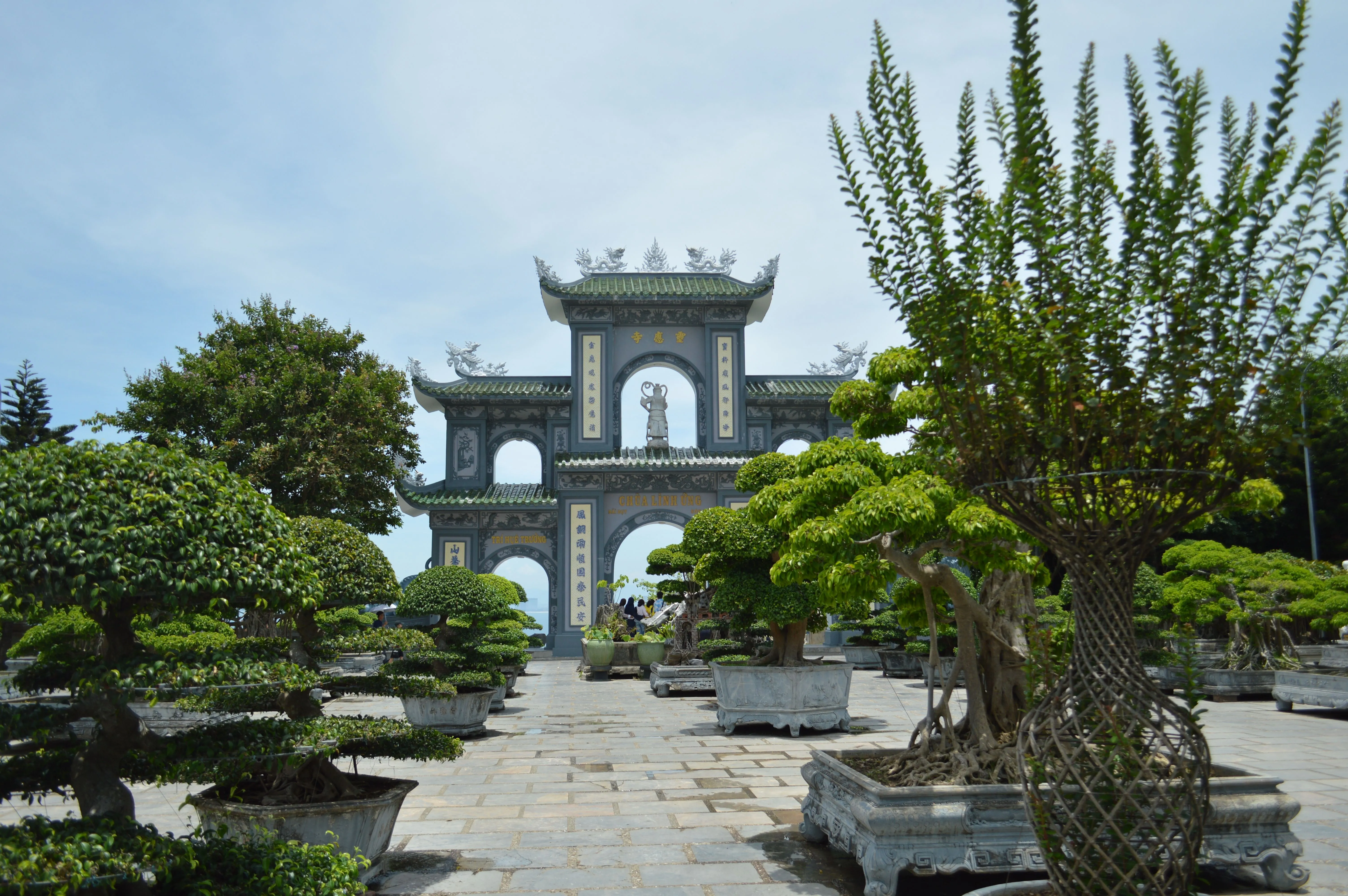 A serene view of a traditional Vietnamese pagoda entrance surrounded by lush bonsai trees in a garden.