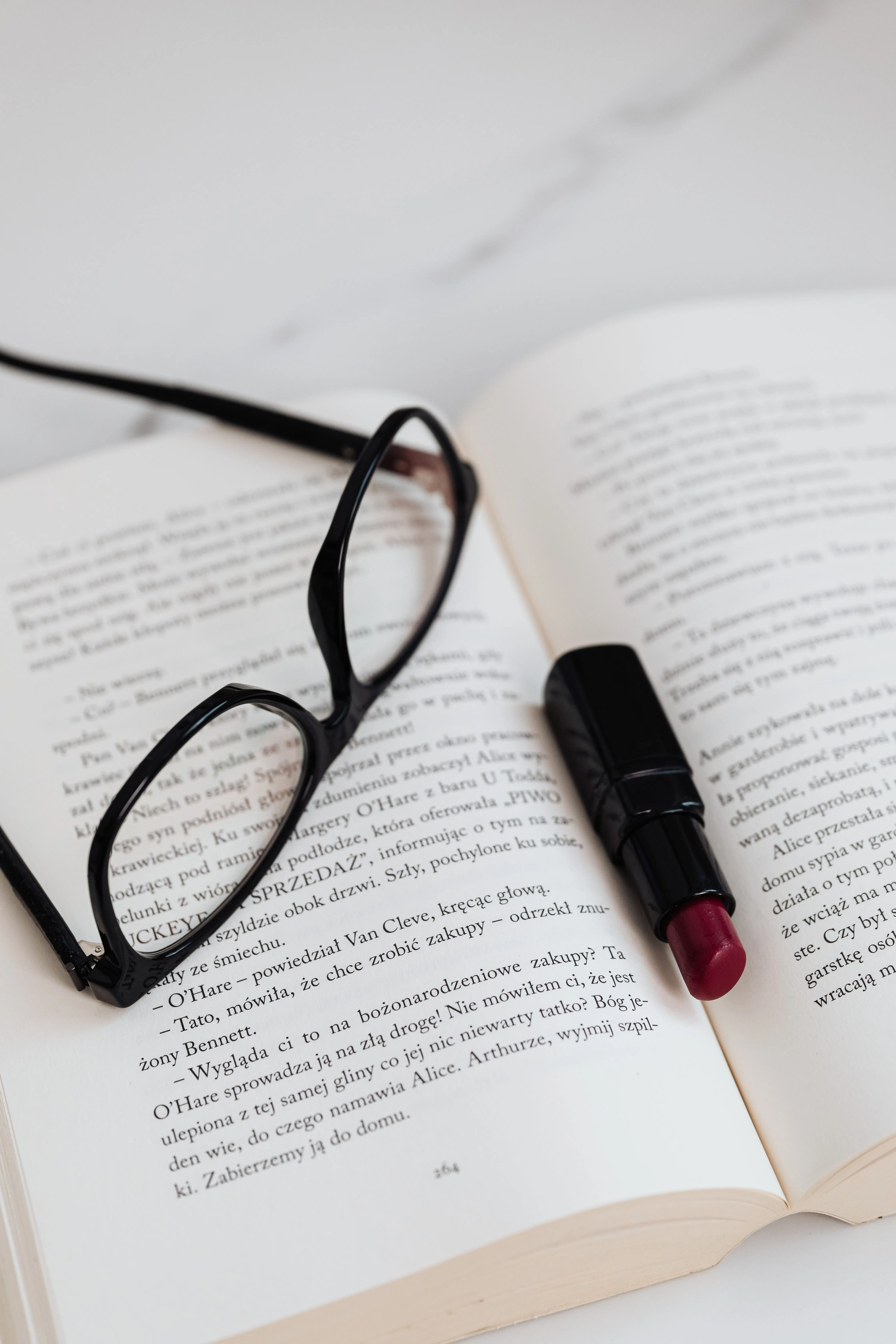A close-up of eyeglasses and lipstick resting on an open book, suggesting a sophisticated lifestyle.