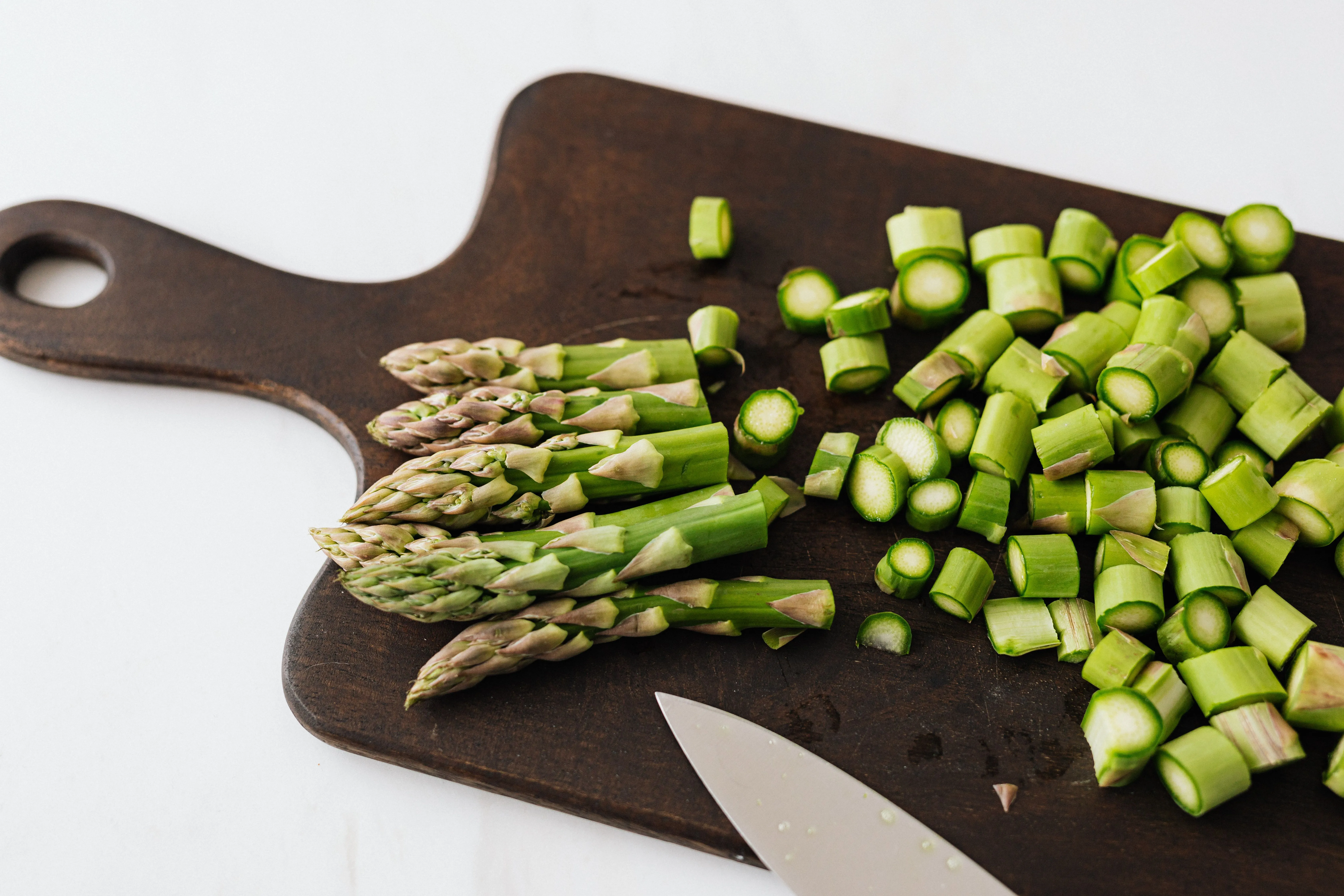 From above of knife near sliced green stems of asparagus composed on wooden cutting board placed on white table