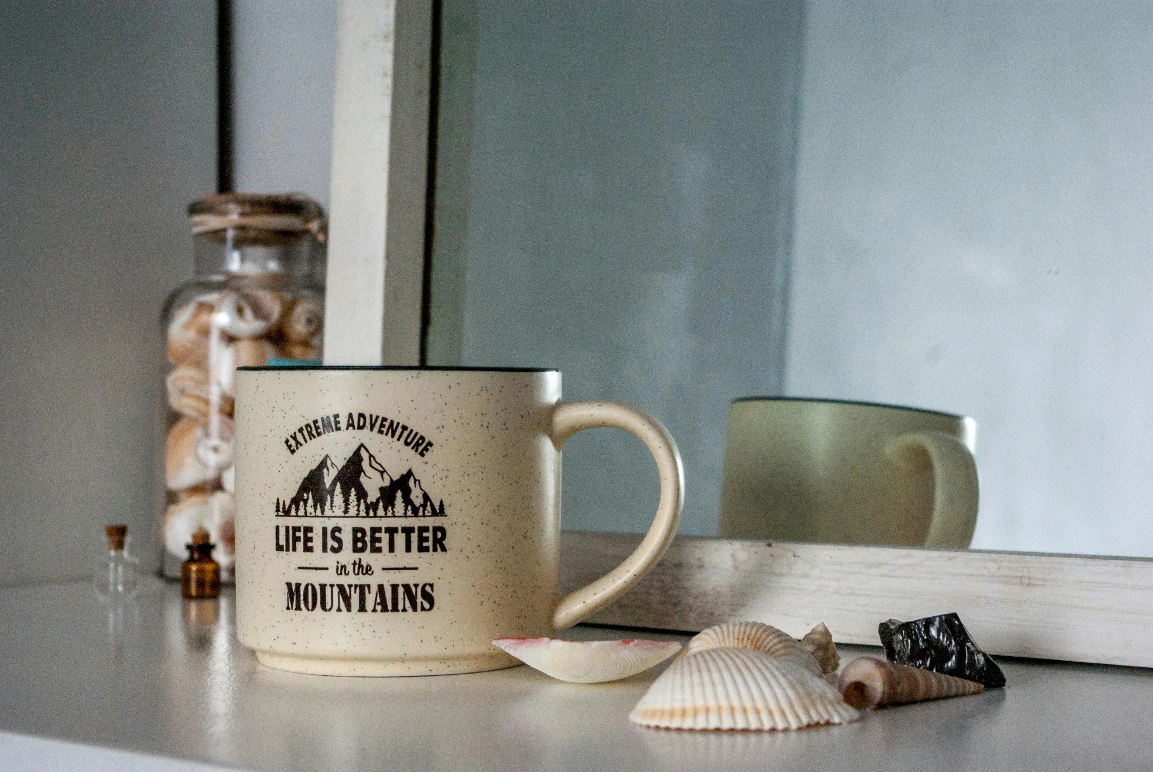 Ceramic light cup with inscription Life is better in the mountains placed near seashells and glass jars on white surface against mirror