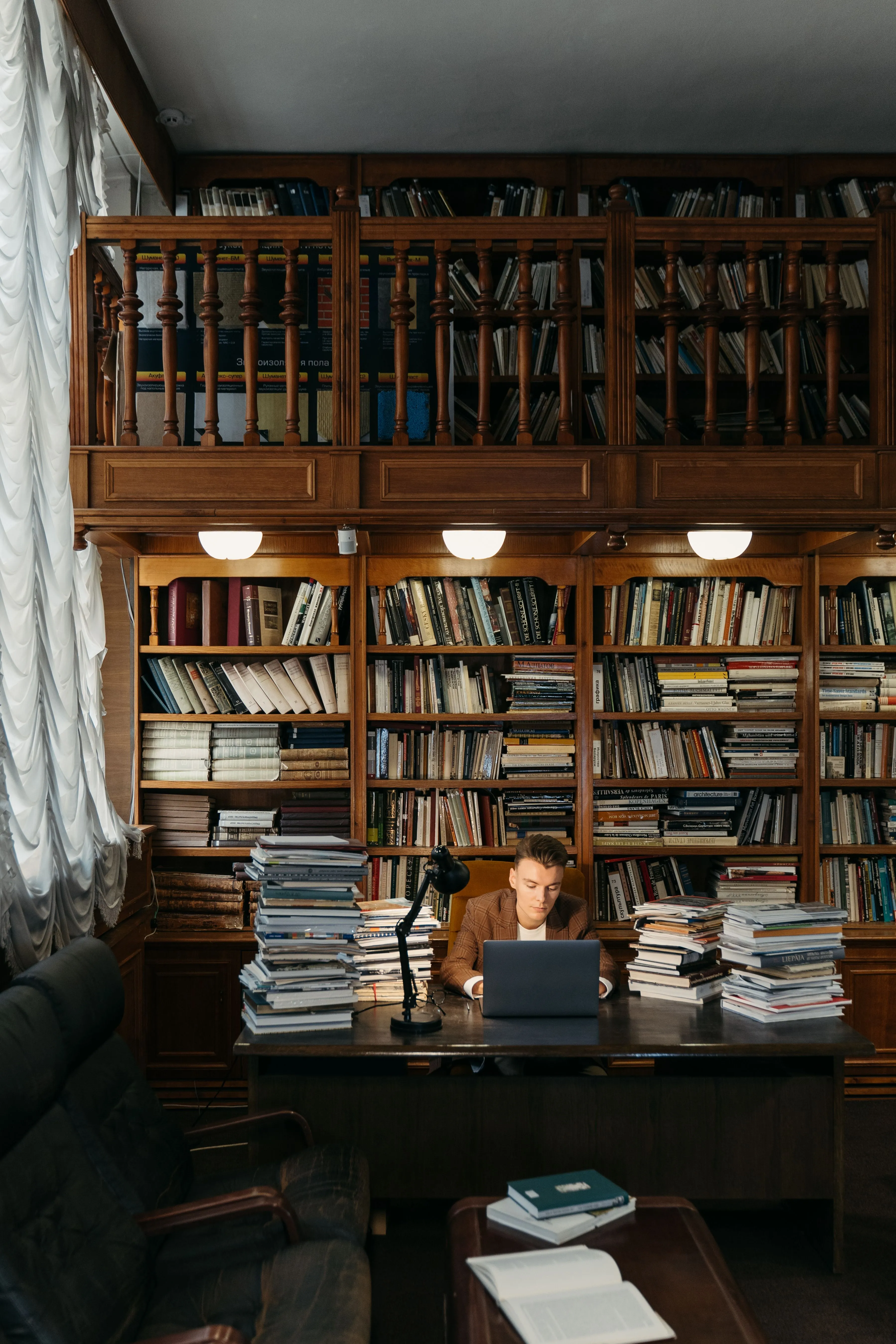 A man sits in a library surrounded by books, reading at a desk with warm lighting.