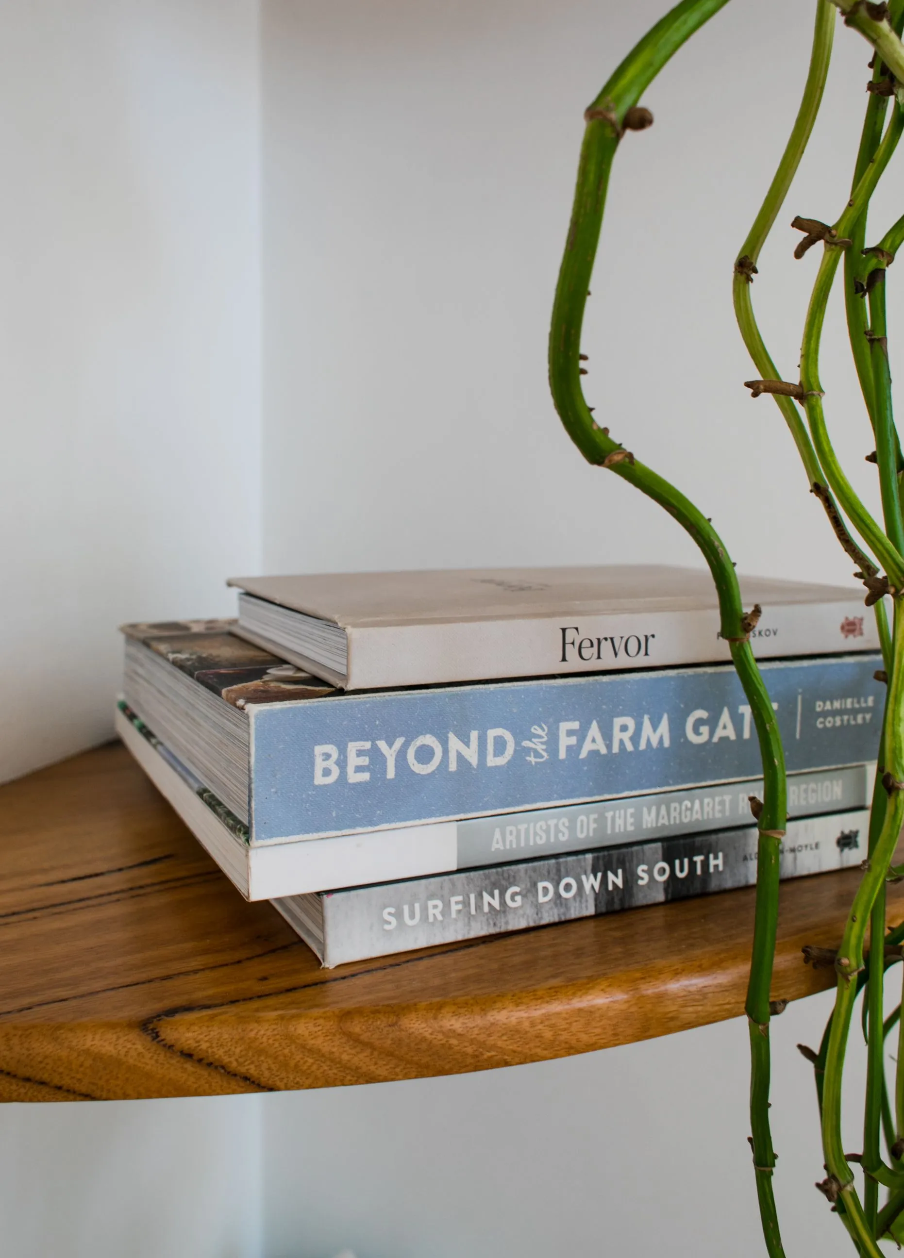 Pile of books placed on timber shelf near fresh verdant branches of plant in corner