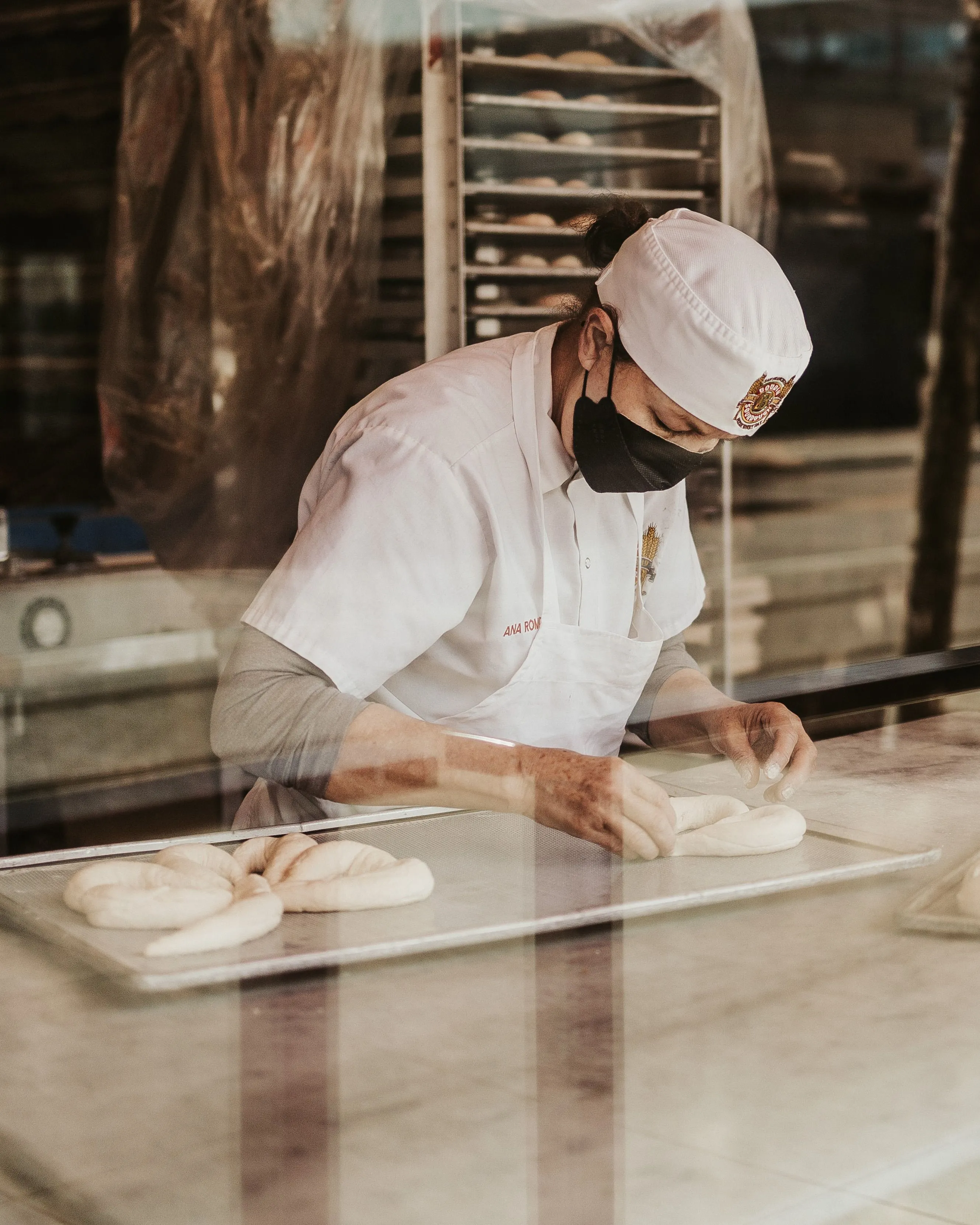 A baker in a white coat and face mask prepares dough in a bakery kitchen.